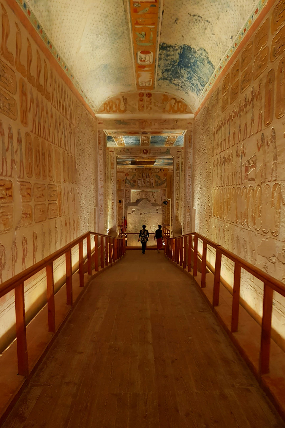 A wooden pathway lined with railing leads into the tombs inside the Valley of the Kings in Luxor. The walls are entirely decorated with hieroglyphs and the passage is long and narrow. 