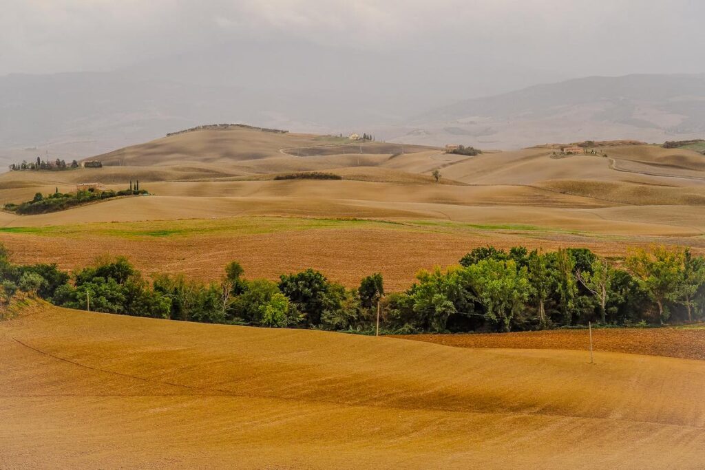 Tuscan hills panorama outside Pienza, featuring serene beauty of Italian countryside under moody sky. Rolling agricultural landscapes in shades of gold and brown with dotted greenery symbolize Tuscany's iconic scenery.