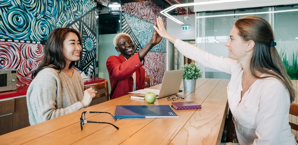 example of coworking space, three people sharing table, two of which are connecting with a high five 