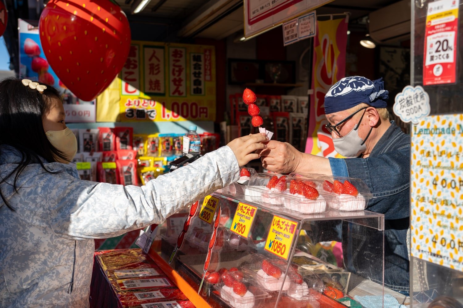Japanese street food vendor handing strawberry candy on a stick to customer with boxed strawberry mochi (ichigo daifuku) on display.