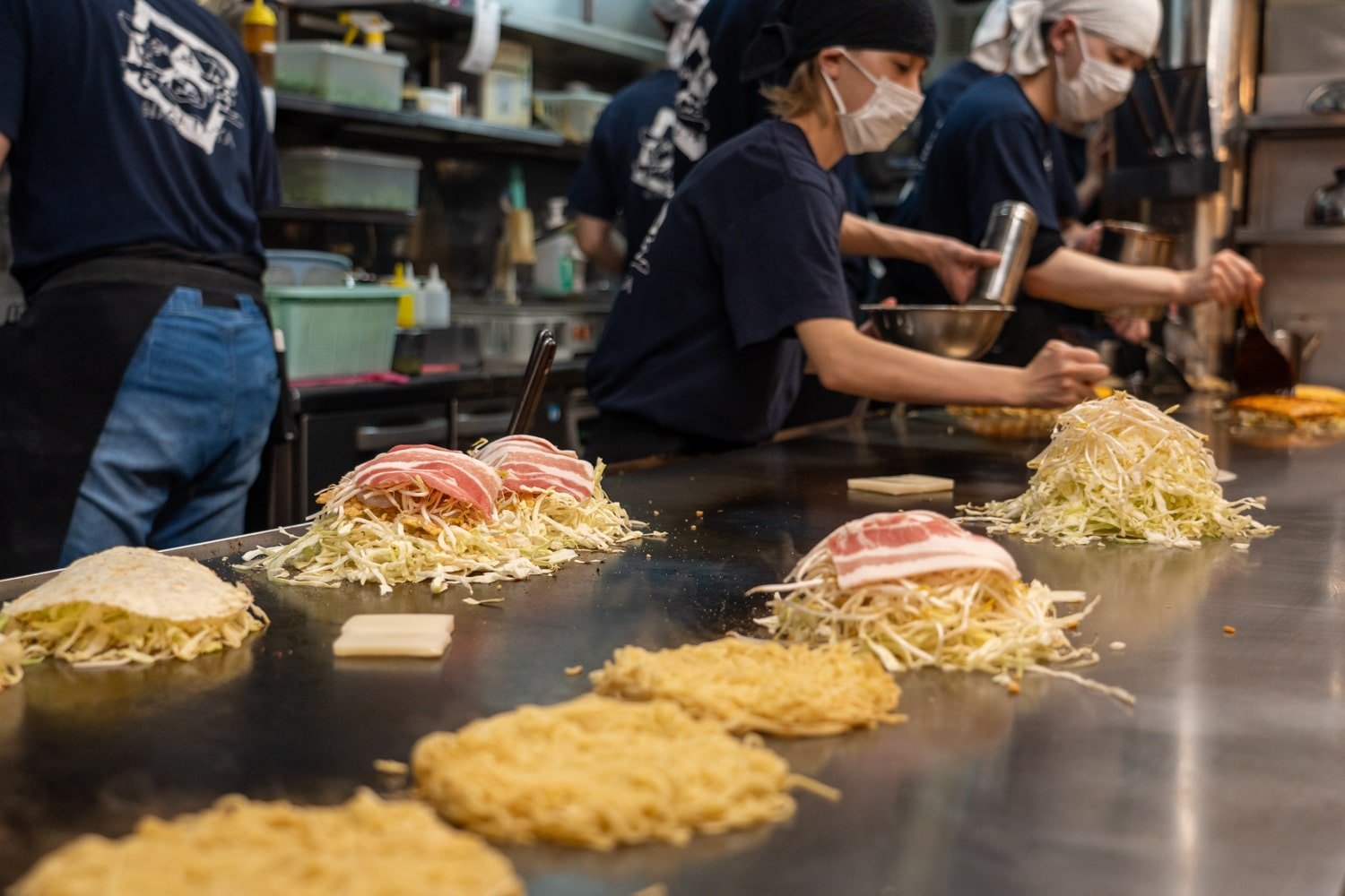 Chefs preparing multiple Hiroshima-style okonomiyaki (Japanese savory pancake) on a hot grill with noodles, eggs, and meats.
