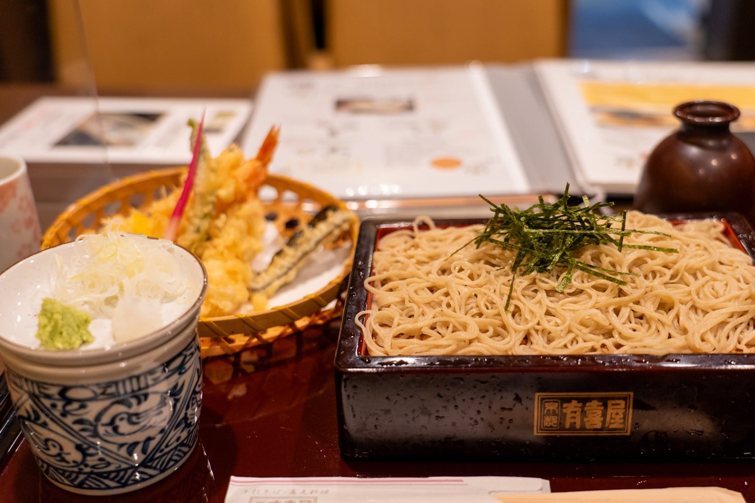 Cold soba noodles set with buckwheat noodles on bamboo tray, a dish of shrimp and vegetable tempura, and a bowl of green onion, dashi broth, and grated ginger.