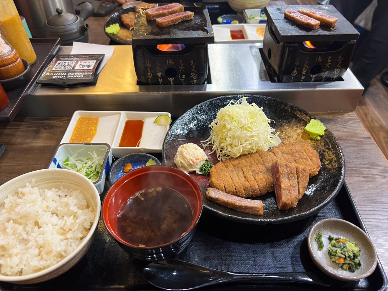 Gyukatsu set (Japanese beef cutlet) with a plate of uncooked, breaded beef cutlet, bowl of rice, dipping sauces, miso soup, and two individual hot plates for grilling gyukatsu.