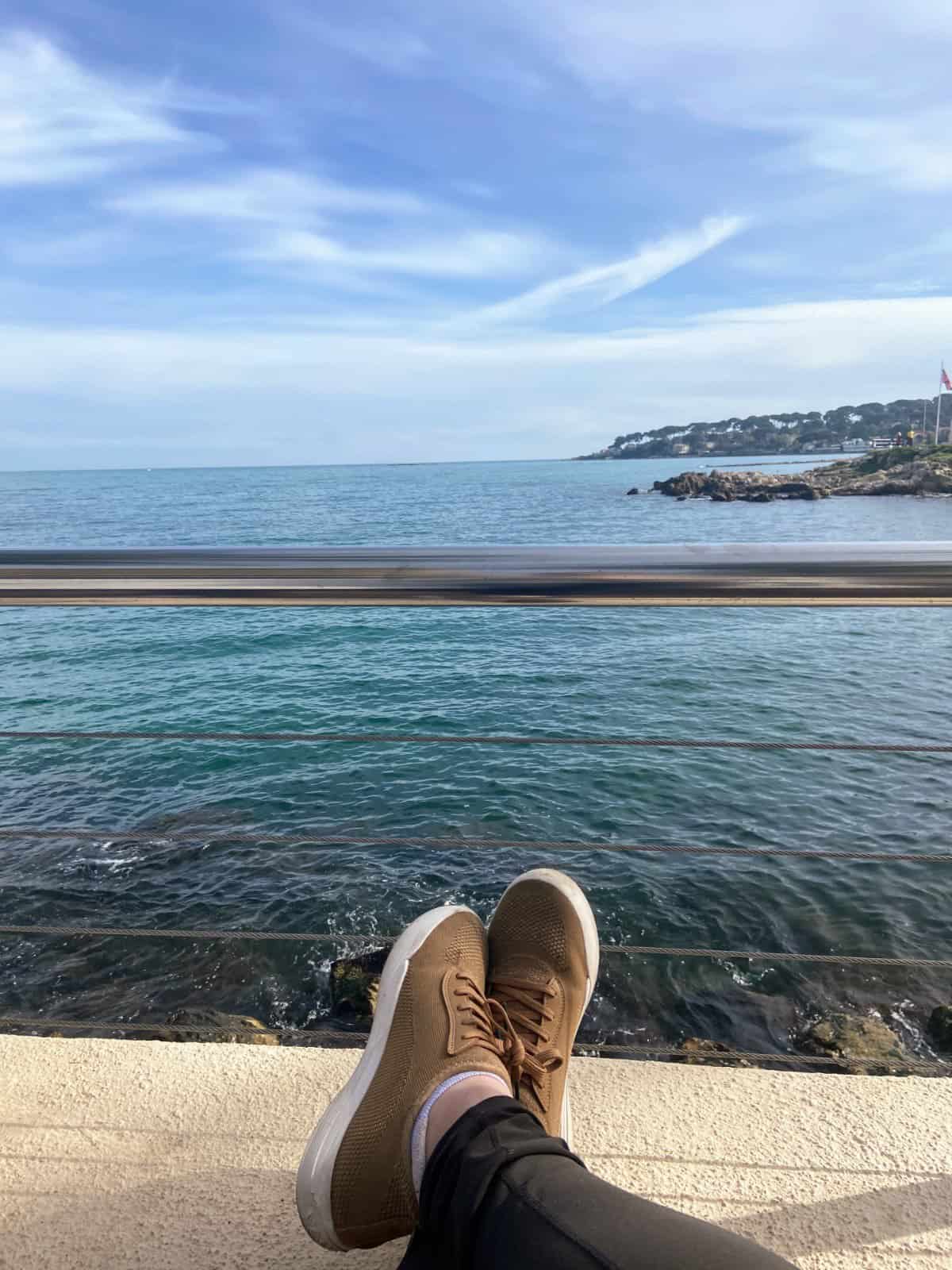 Riana sitting with her ankles crossed and propped up on a wall overlooking the water in Antibes, France showing off her Vessi shoes 