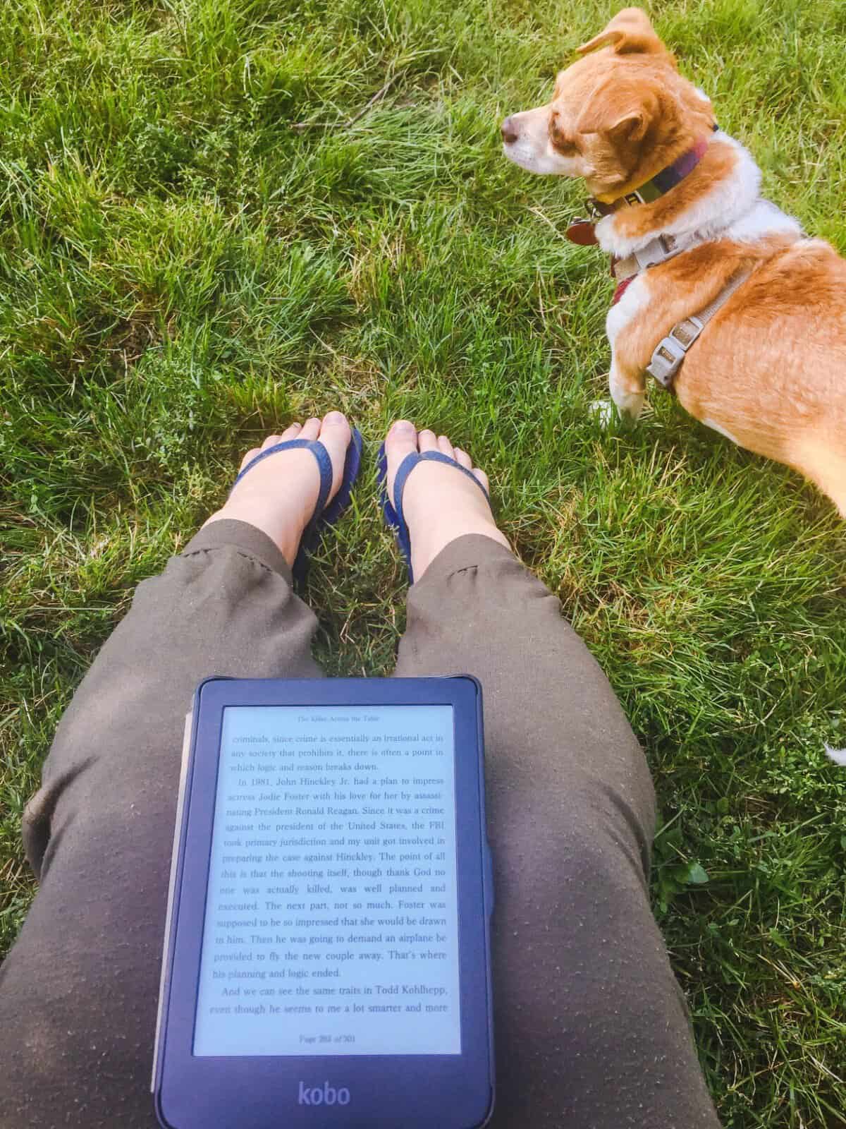 Shot of Riana's legs with an ebook in her lap and her dog Ellie on the grass in front of her