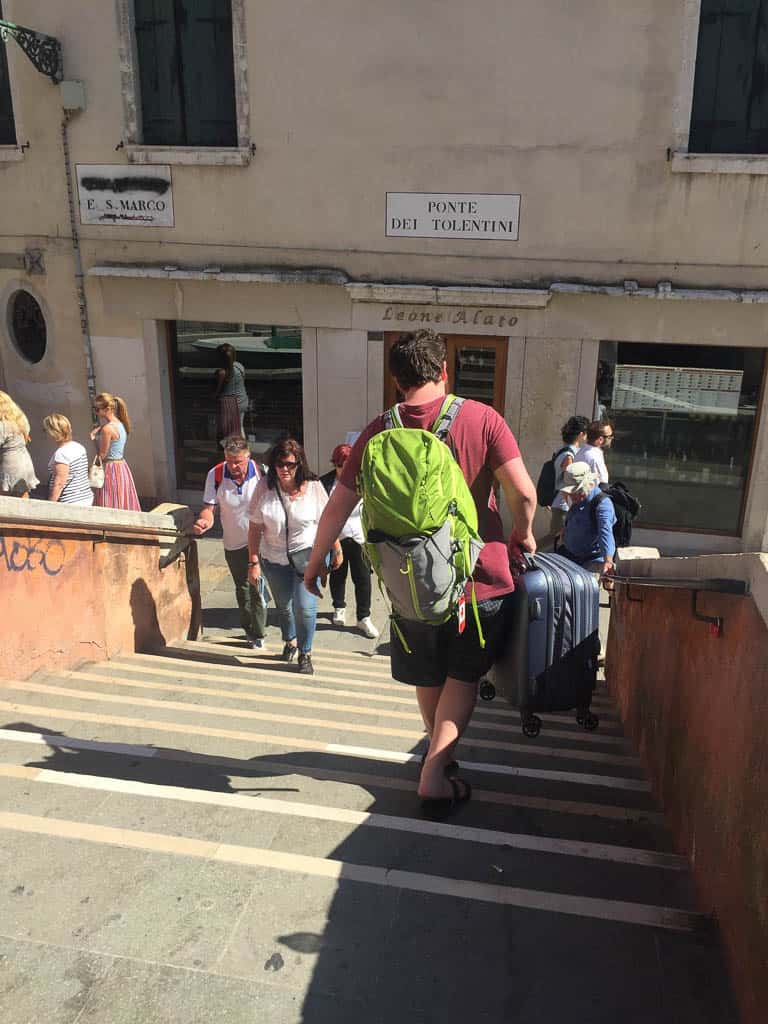 Colin wearing a backpack while carrying a suitcase down the steps of a Venice canal bridge