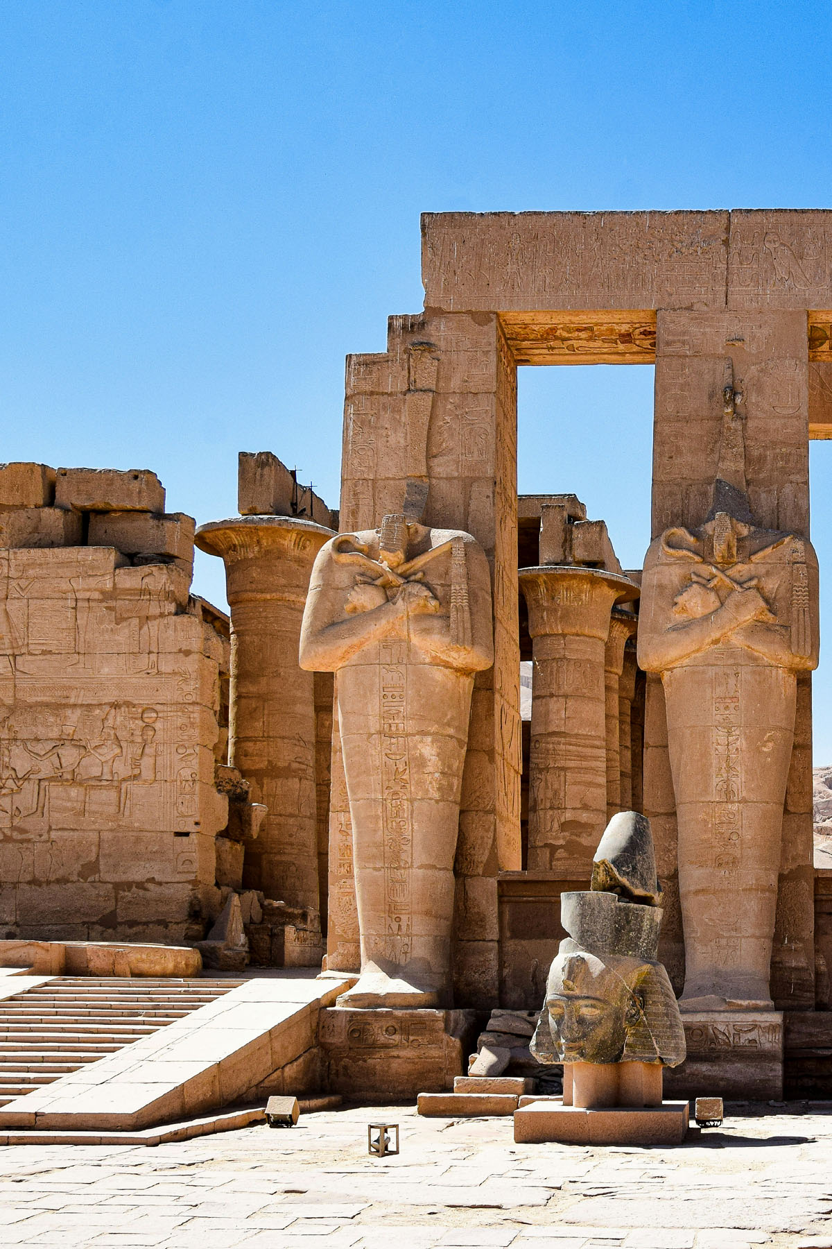 A fallen head of a statue of Ramses II lays on the ground alongside a row of standing statues at the Ramasseum in Luxor.