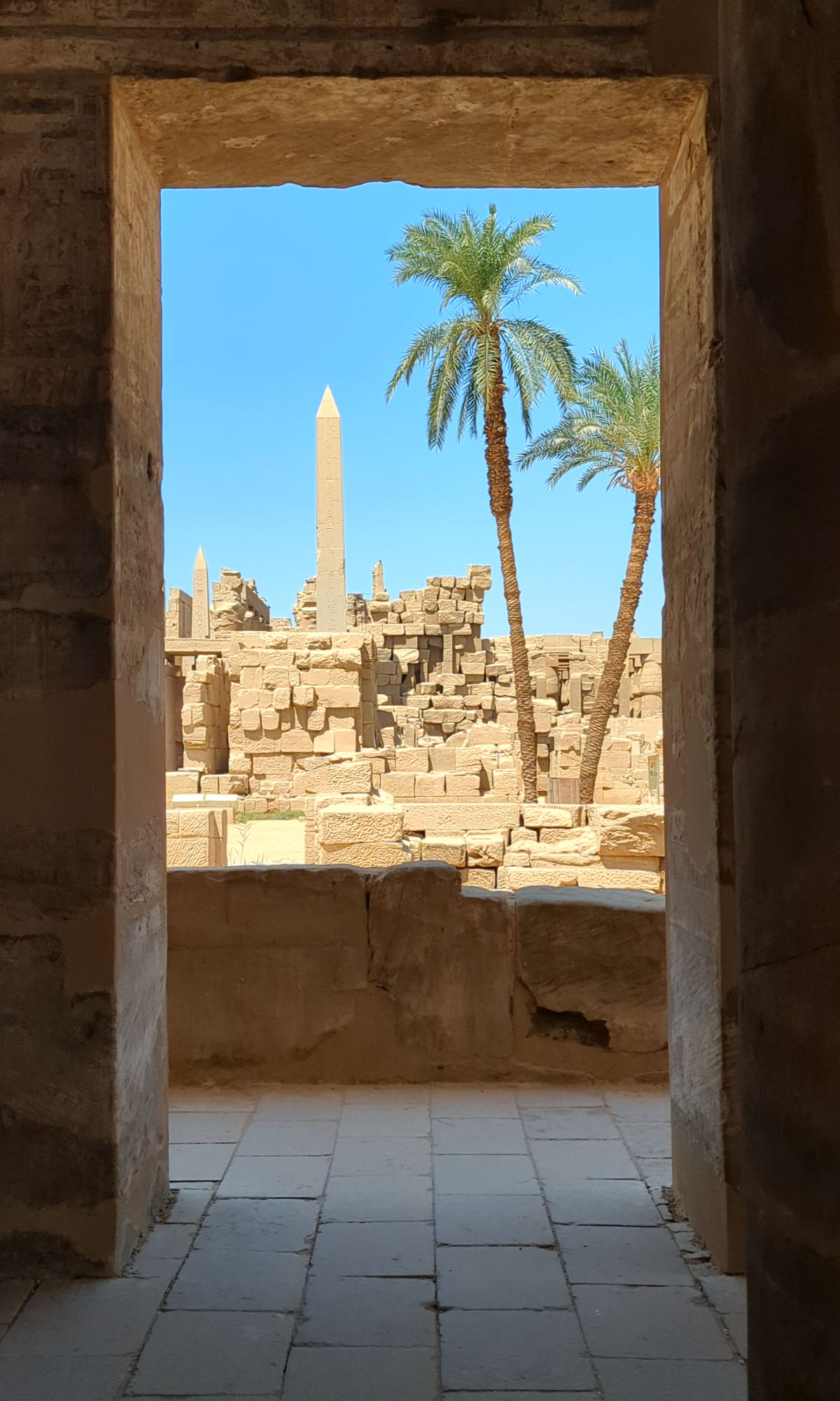 An obelisk rises up against a blue sky with a square dark entrance in the foreground at the Karnak Temple in Luxor. 