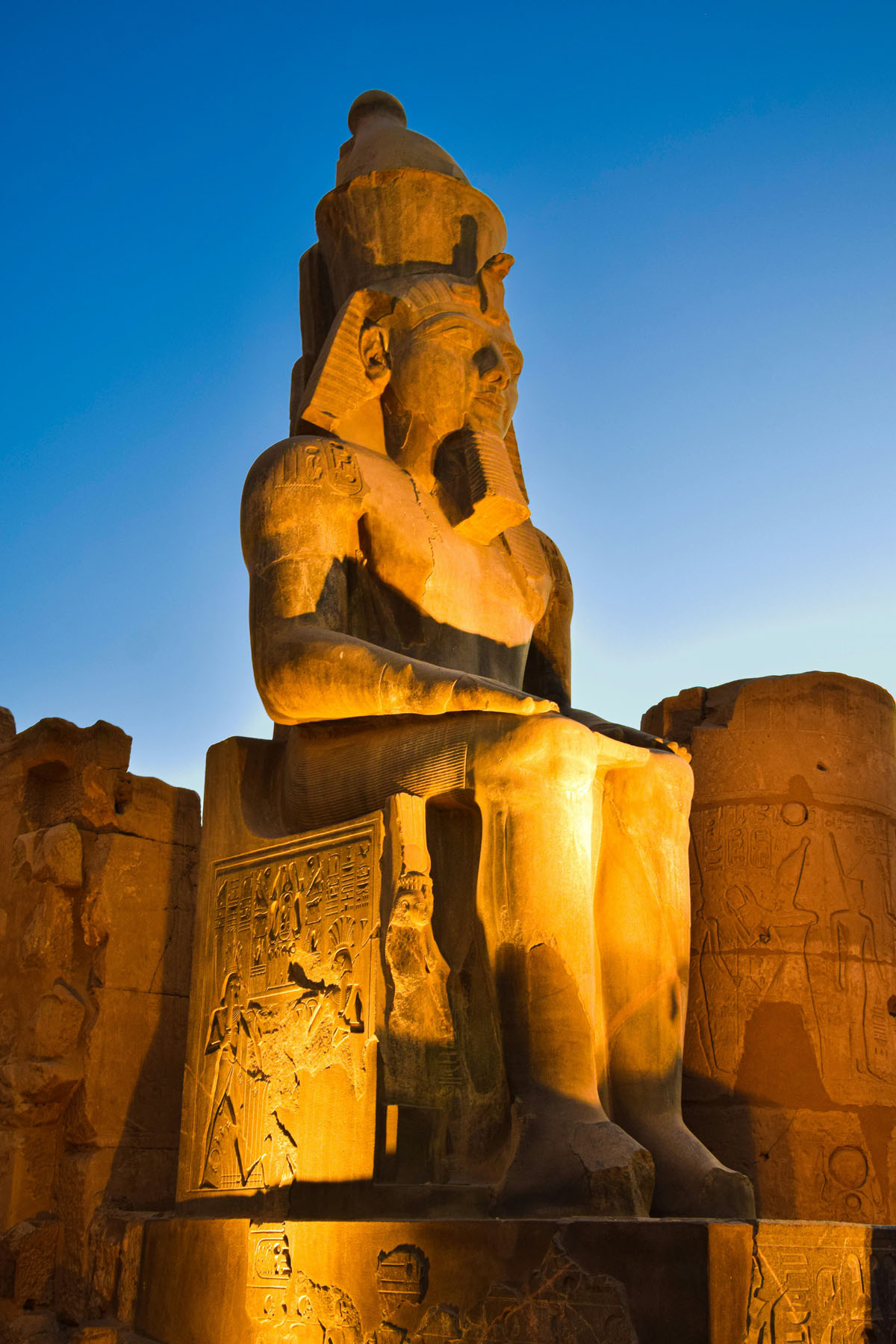 An enormous statue of Ramses II at the Luxor Temple in Egypt lit up at night in golden light. 