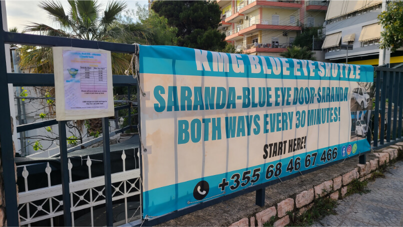 Bright blue and white sign for shuttles to Blue Eye. Sign reads: Saranda to Blue Eye, both ways every 30 minutes.