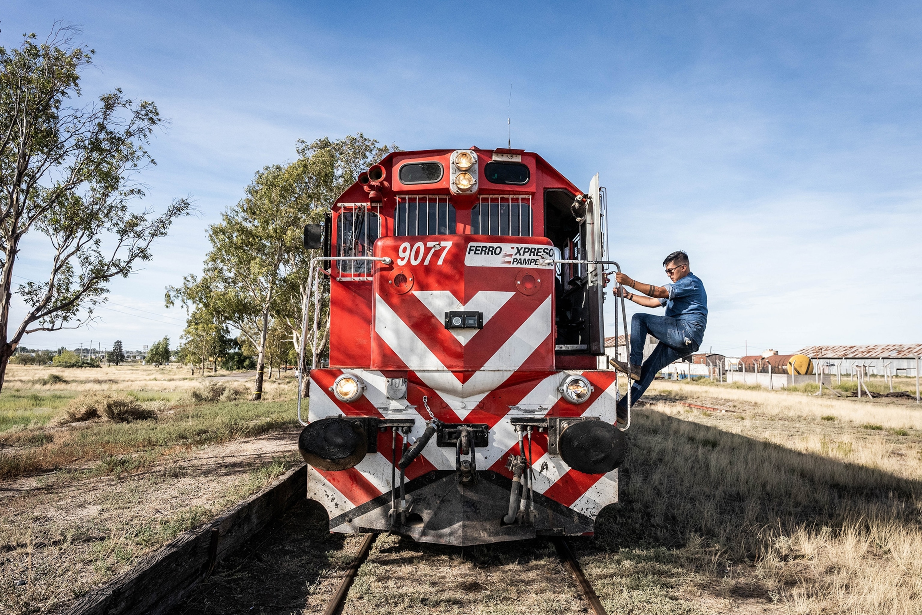 A man clad in denim shades, climbs onto the front red and white train surrounded by short brush