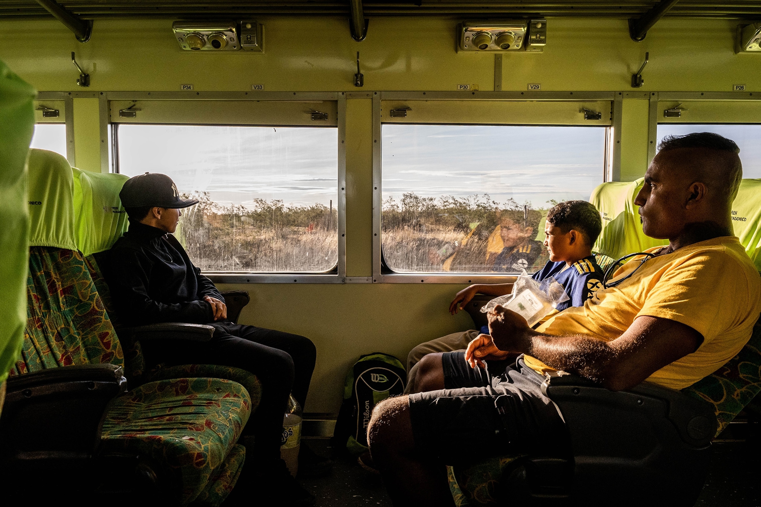 A young boy inn a Yankees hat anothe rin a blue and yellow jersey sit with a man in a yellw shirt warm sunlight spilling into the right side of the train the boy in the yellow squints as they all look at the scenery.