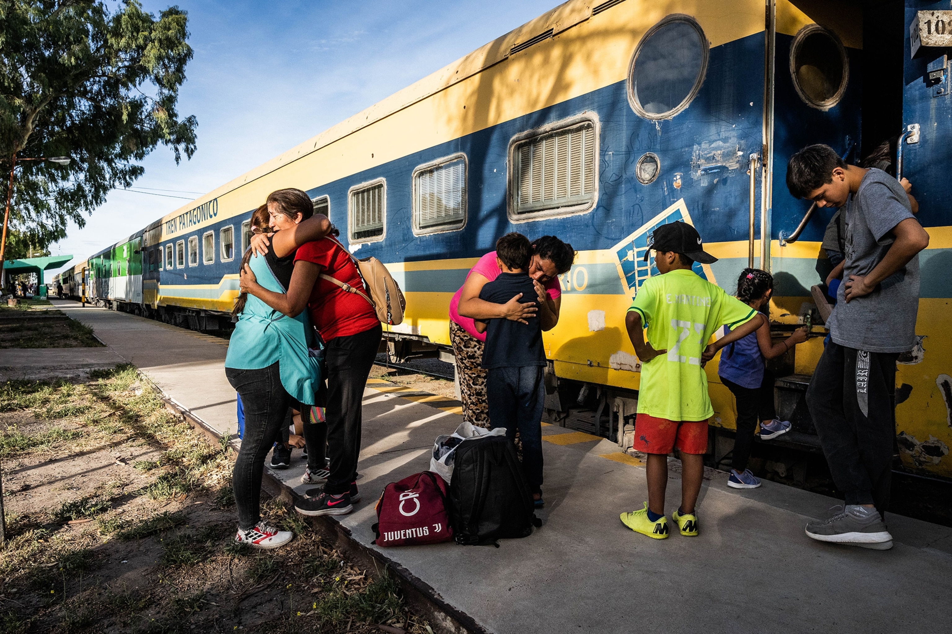 A family embraces each other in front of the yellow and blue train clear blue skys and the worm glow of the sunlight illuminating them.