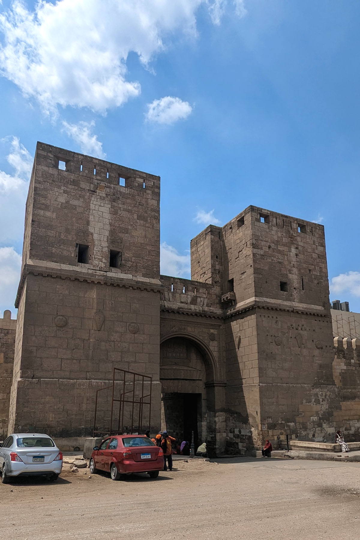The medieval gate of Bab al-Nasr stands on a street with a few packed cars in Cairo. It has huge square towers that flank an ornate arch.