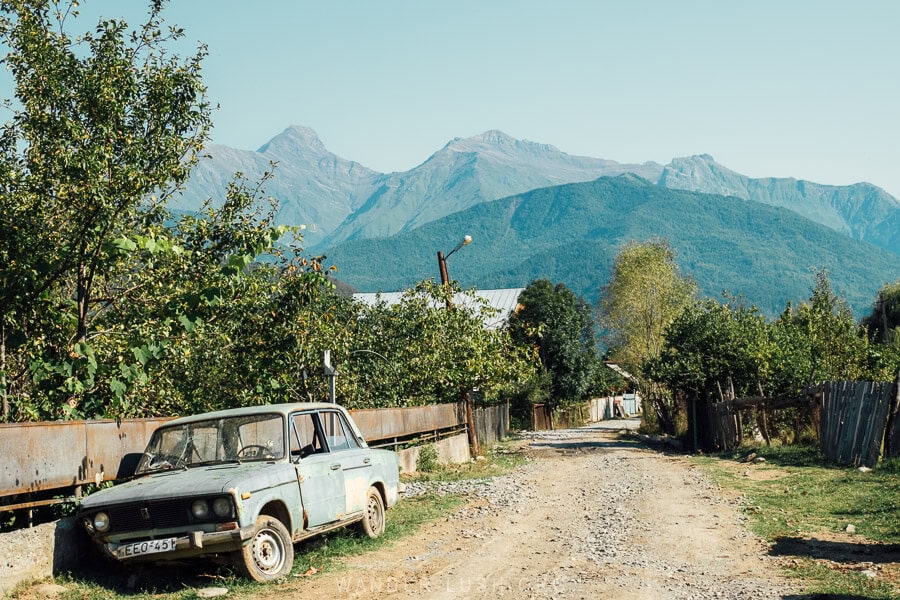 An old car parked on a rural road in Racha, Georgia with a backdrop of beautiful mountains.