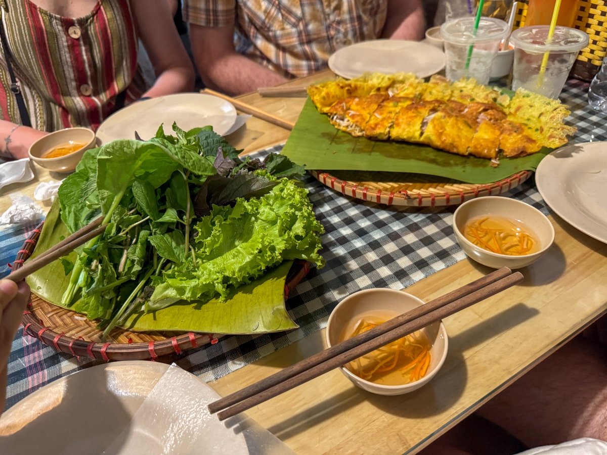 Table with Vietnamese food—banh xeo—sizzling fried savory pancake with plate of herbs and chopsticks.