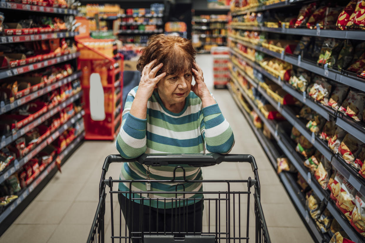 Senior woman standing among the produce aisle at the supermarket and feeling worried about the increase in groceries prices