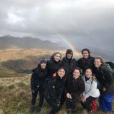 Rosa with friends on top of a mountain of beige/green grass fields. There are grey storm clouds in the background and rain in the distance. Sun shines through a hole in the clouds and a rainbow arches out behind the group. The group is primarily wearing black overcoats.