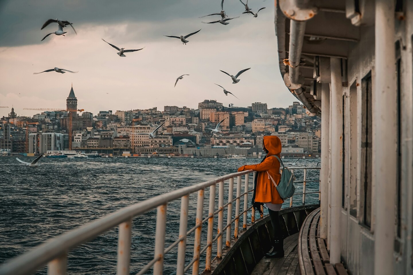 Person in an orange hooded jacket standing on a boat with the Istanbul skyline in the background and seagulls flying overhead