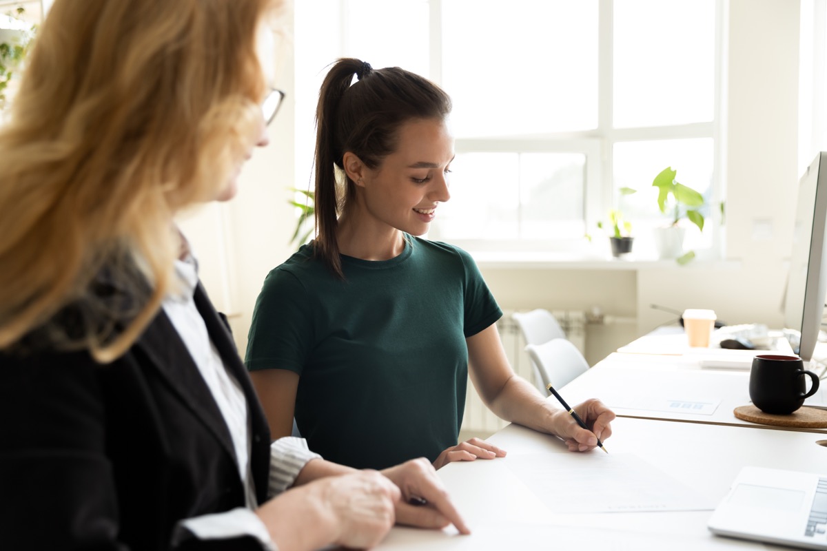 Mature business teacher supervising happy student writing notes. Middle aged mentor training intern at office workplace. HR manager helping new employee to fill and sign employment agreement