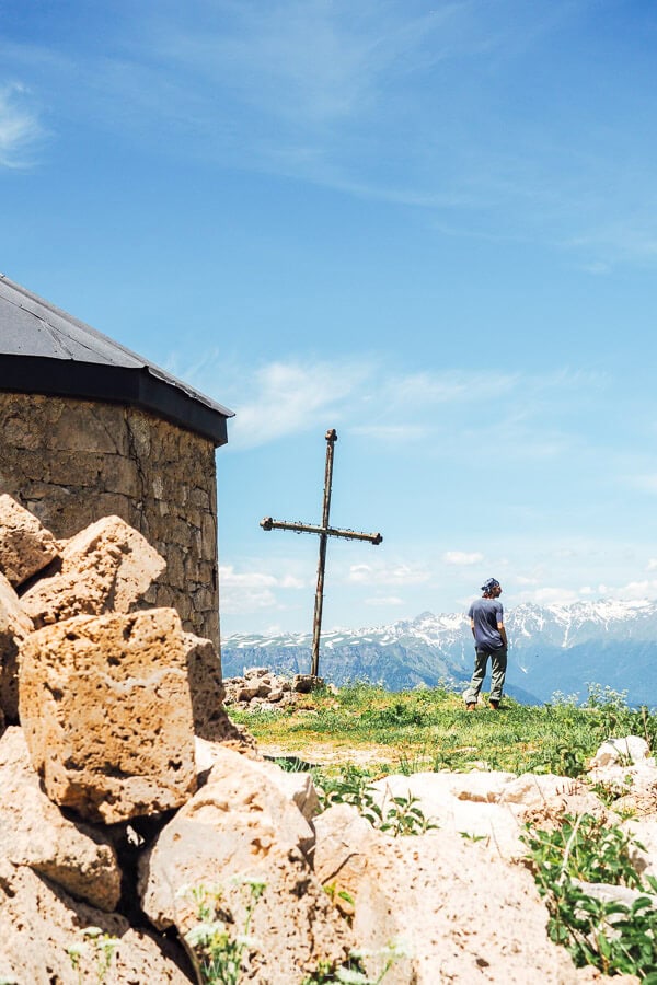 A man standing near a stone church and cross on a plateau overlooking the Caucasus mountains.