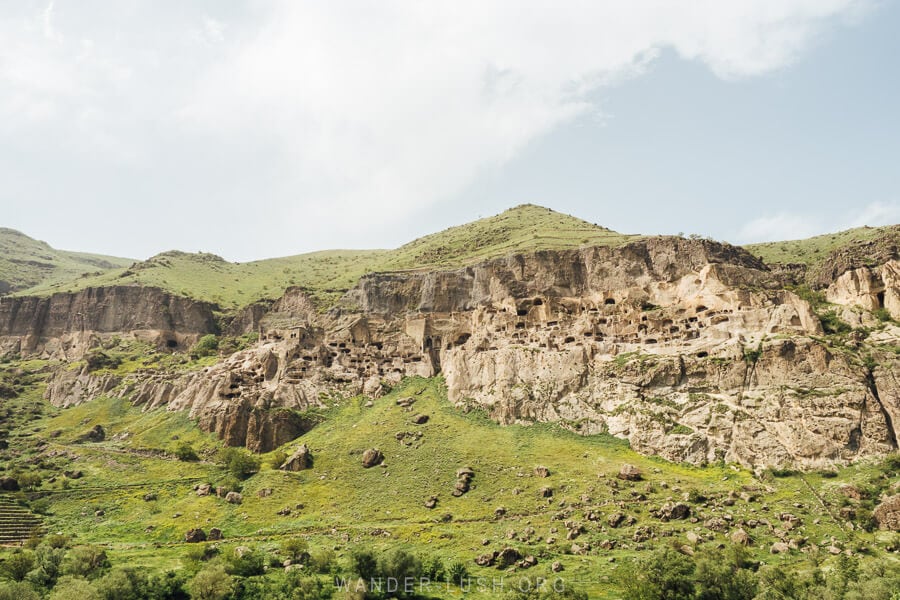 A view of Vardzia Cave Town, with stone chambers cut from the rock face and green grass all around.