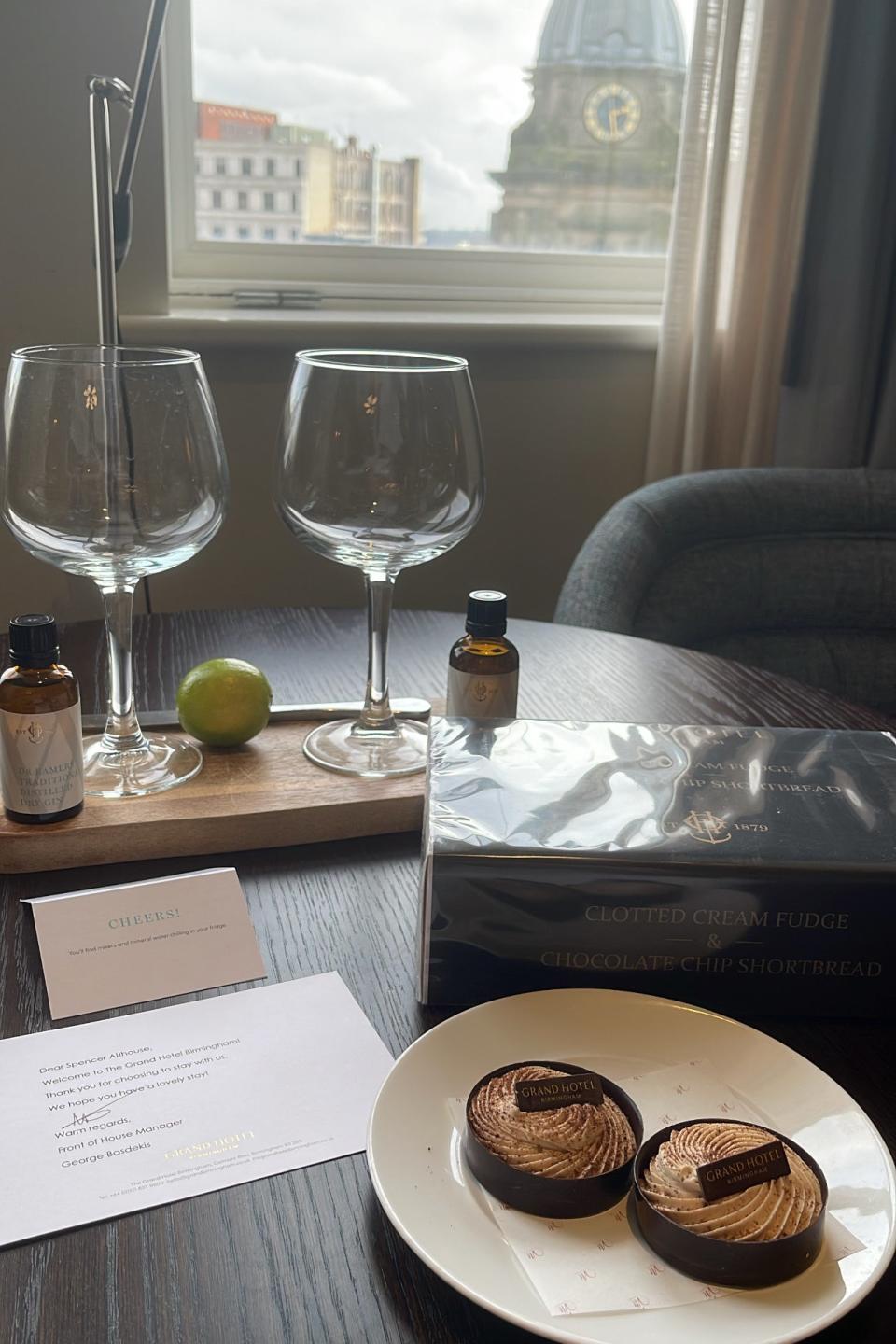 Table with wine glasses, small bottles, a lime, a note, chocolate fudge and chocolate chip shortbread near a window. Cityscape with a dome in the background