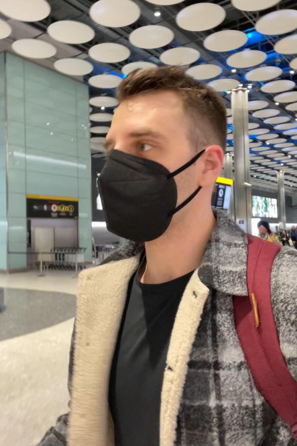 A man wearing a black mask and a plaid coat with a woolen collar walks through a modern indoor space with patterned ceiling lights