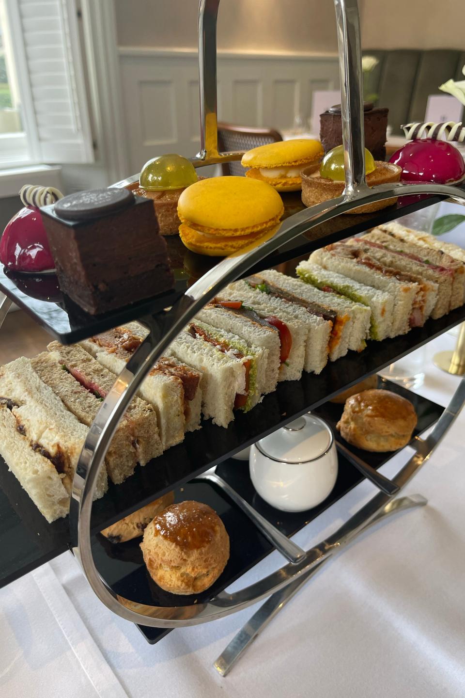 Two-tiered afternoon tea tray with assorted finger sandwiches on the lower tier, and various sweets, including macarons and pastries, on the upper tier