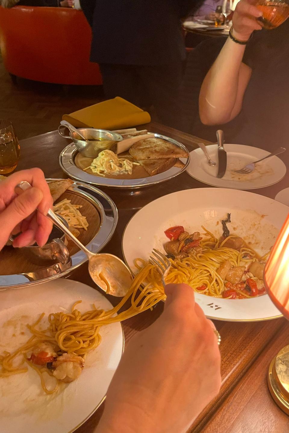 Two people eating a pasta dish at a dimly-lit restaurant. A person's hand is twirling spaghetti on a fork, and there are various other plates of food on the table