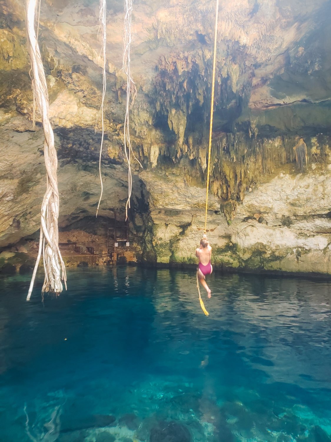 Tourist swinging on rope into beautiful blue, semi-open cenote in the jungle (Cenote Yaxbacaltun) near Cuzumá and Homún, Yucatán, Mexico.