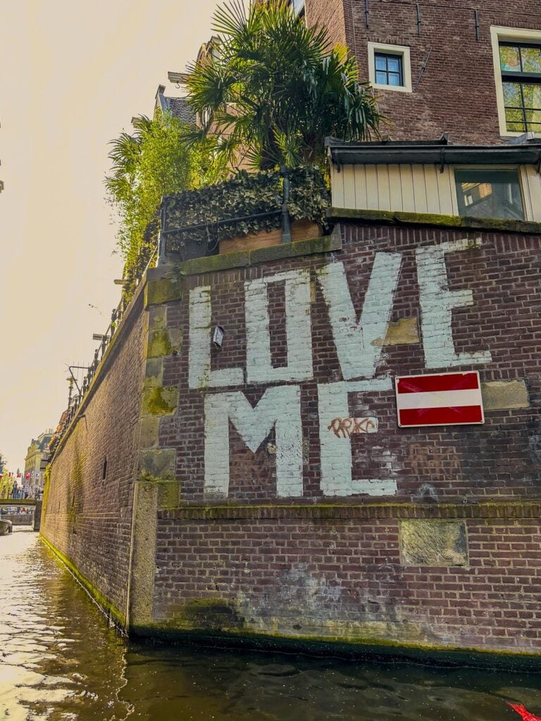 Love Me street art on historic canal-side brick wall in Amsterdam with lush greenery and red and white sign.