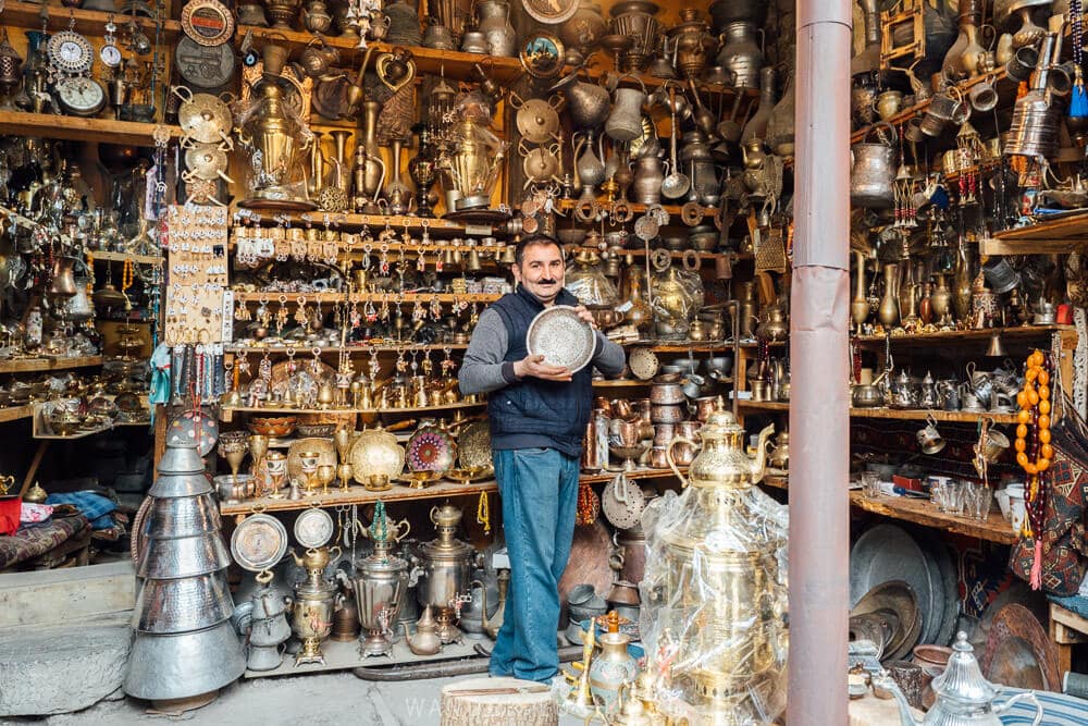 A man holds a metal plate in front of a shelf overflowing with urns, jugs and other items at a workshop in Azerbaijan.