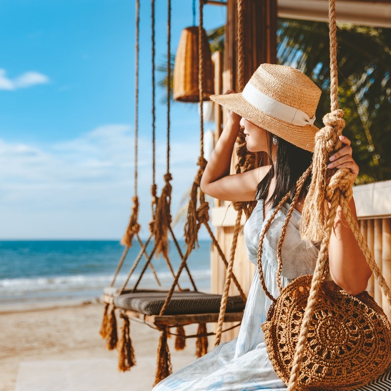 Young Female Traveler Wearing A Straw Hat And A Summery Dress As She Sits On A Beach Swing Looking Out To The Ocean, Beach Vacations