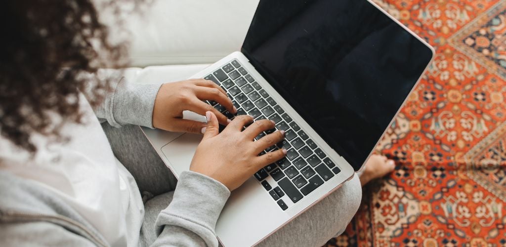 woman typing on laptop, sitting on sofa, colorful rug beneath feet 