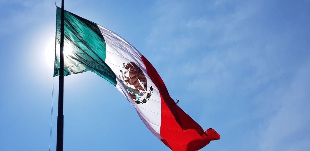 Mexican flag against blue skies and sun rays shining through 