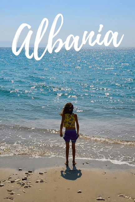 A child stands on a sandy beach in front of the blue water of the sea in Albania.