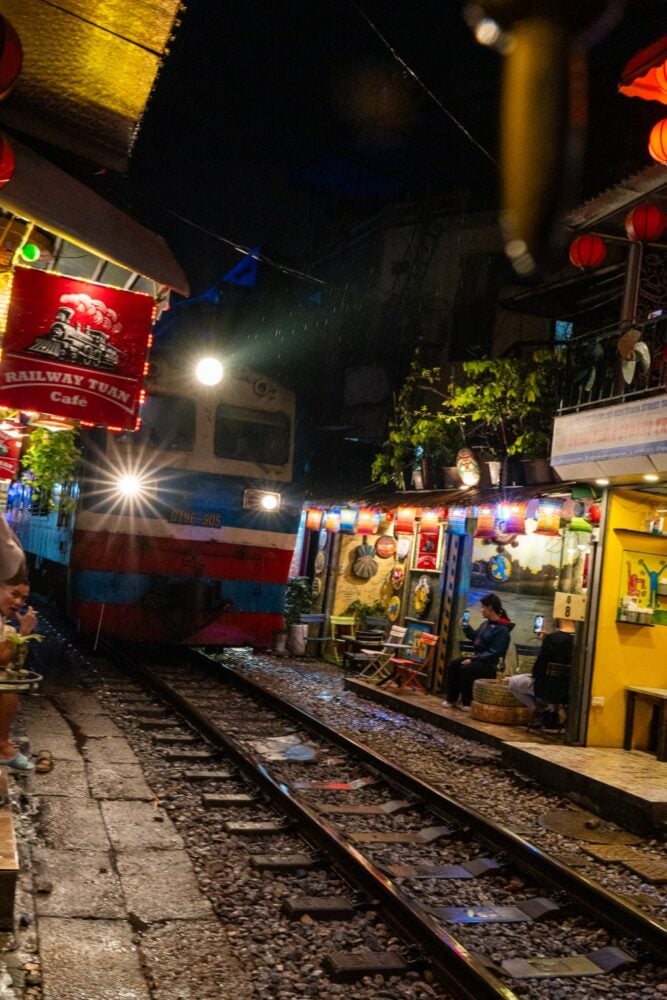 Train arriving on Hanoi Train Street at night with cafes and shops on narrow street.