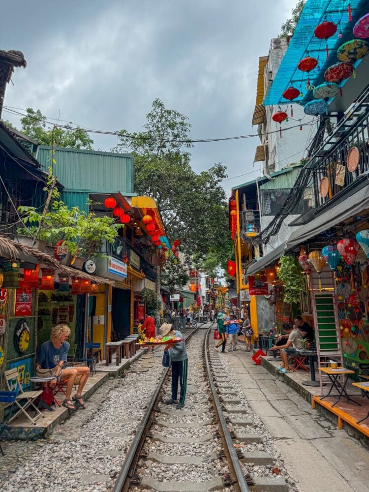 Local Vietnamese woman selling wares on Hanoi Train Street during the day with shops and cafes decorated with lanterns lining the railroad tracks.