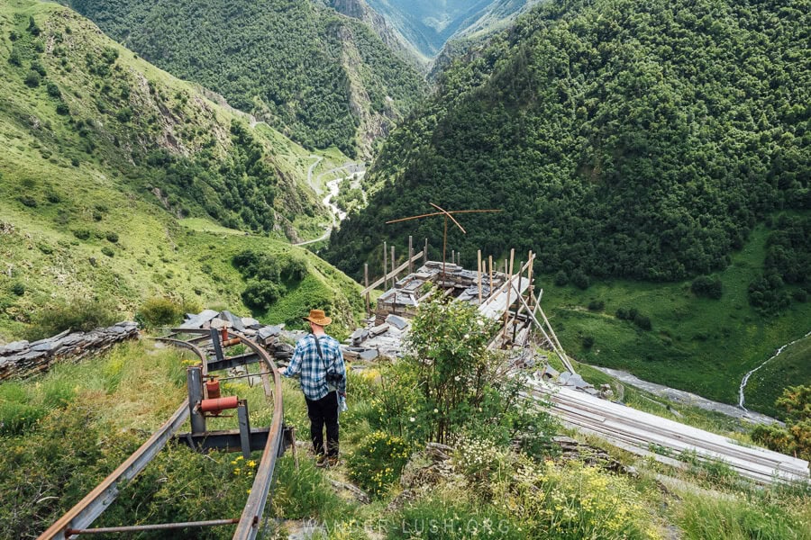 A man walking through the grass above a river valley alongside construction equipment in Mutso, Khevsureti.