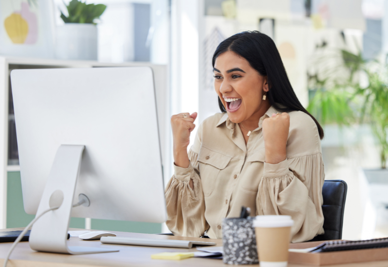 A woman with long, dark hair sits at a desk and celebrates success with raised fists and a joyful expression in front of a computer screen.