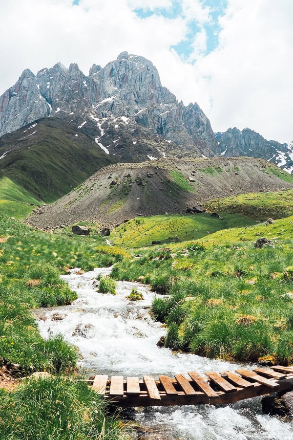 Mount Chaukhi rising above a fast moving river with a small wooden bridge in the Juta Valley.