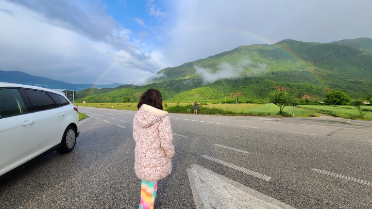 A girl in a puffy jacket stands next to a white rental car beside the highway in the mountains with a full rainbow in the distance.