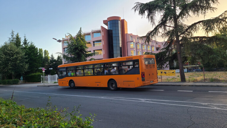 The orange tourist city bus in Sunny Beach as it drives past a pink building with a circular corner.