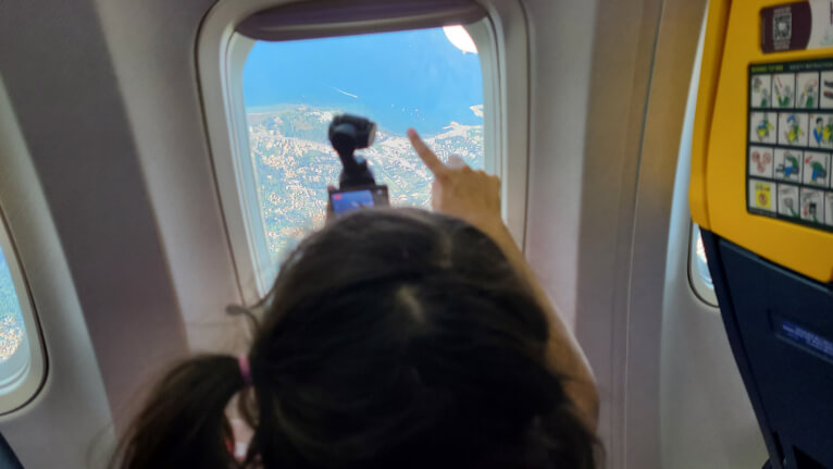 A girl with pigtails holds a camera up to a plane window with a beautiful coastal scene below.