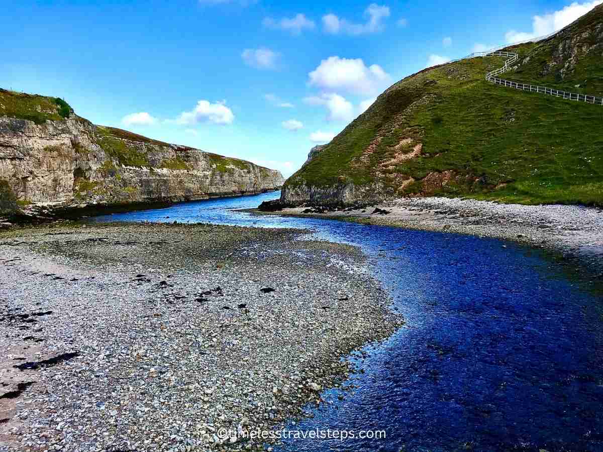 Image of water flowing between the cliffs of Smoo Cave, extending out to the sea. The clear, blue water is bordered by rugged, weathered cliffs, creating a dramatic passage that leads the eye to the open ocean. The scene captures the natural beauty and dynamic interaction between the land and sea in this stunning coastal landscape.
