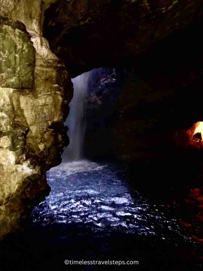 Image of the Smoo Cave waterfall with water gushing through a natural opening in the cavern's roof, cascading down into the pool below. The powerful flow of water creates a dynamic scene, with mist rising and light filtering through the opening, highlighting the cave's rugged rock formations and adding to the dramatic atmosphere.
