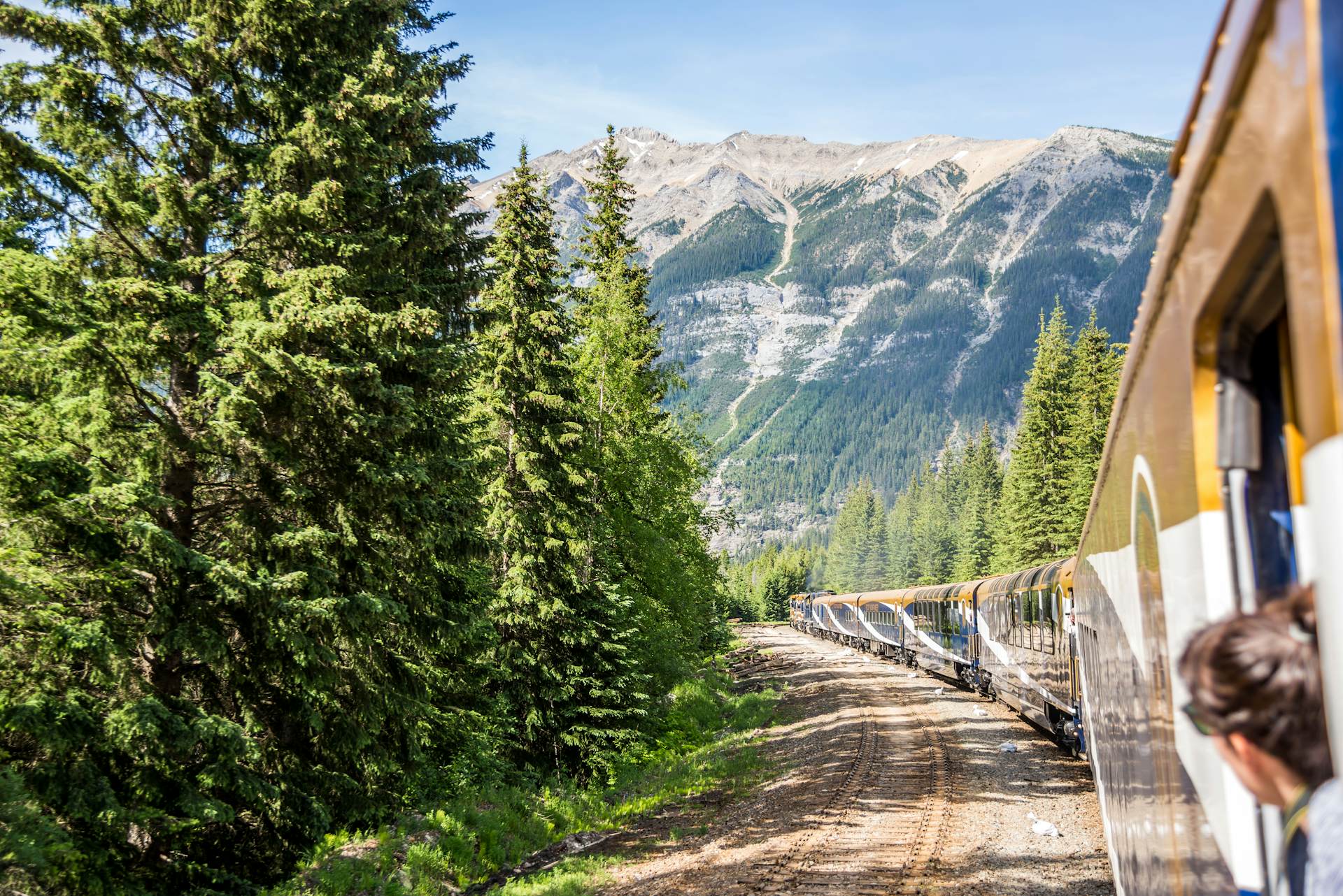 A person nudges their head out of a train window in an open carriage to see the mountain scenery