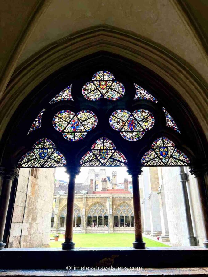 Image of the cloisters at Westminster Abbey, featuring an arched window with stained glass at the top, overlooking the tranquil quad. The Gothic arch and the vibrant stained glass add to the serene and contemplative ambiance.
