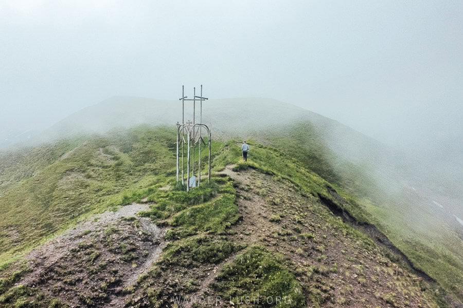 A man walking along a misty ridge in front of a cross sculpture on the Datvisjvari Pass.